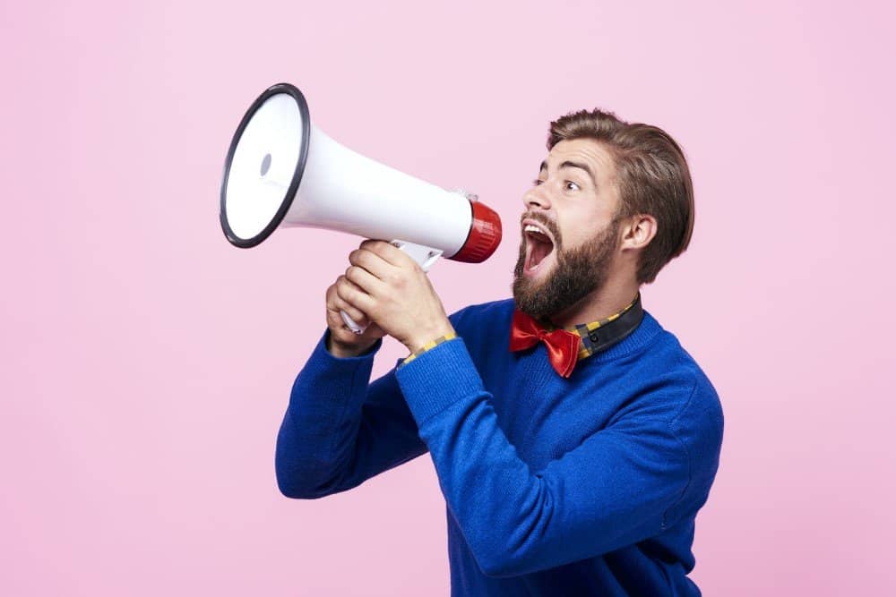 A bearded man shouting into a megaphone on a pink background, embracing Loud Budgeting.
