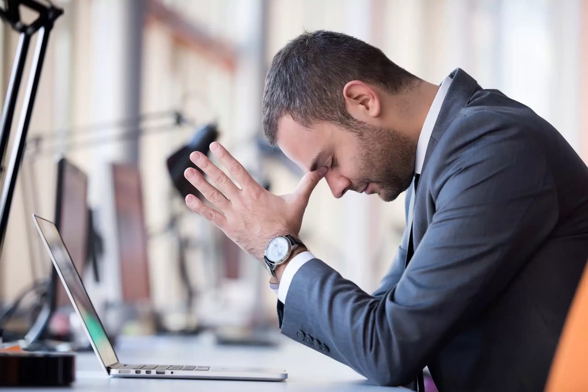 A man in a suit is holding his head in front of his laptop.