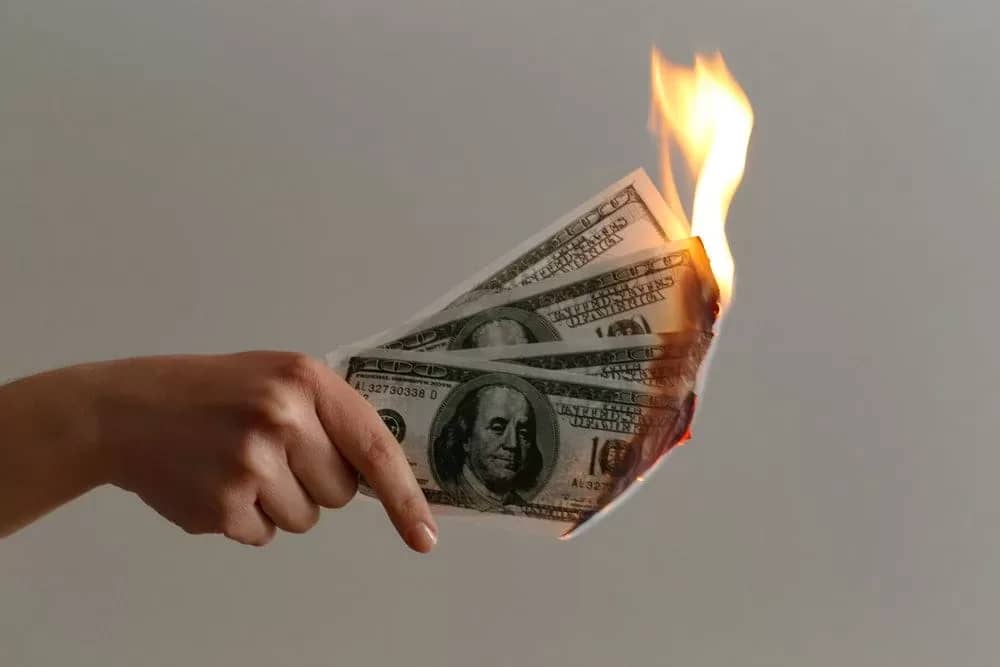 A hand is holding a burning dollar bill, symbolizing the end of bad money habits.